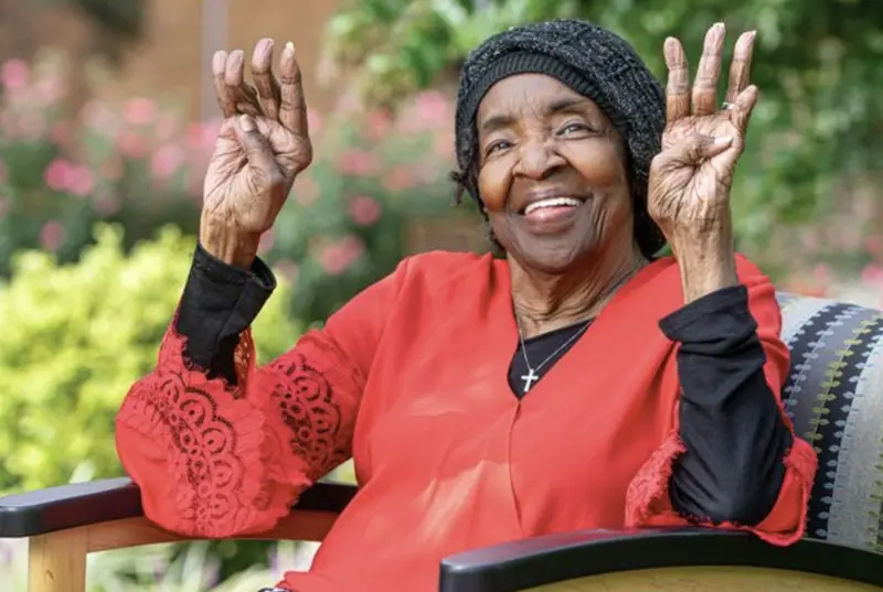 Ms. Alice “Bunnie” Boswell, smiling with both hands raised, seated outdoors among flowers
