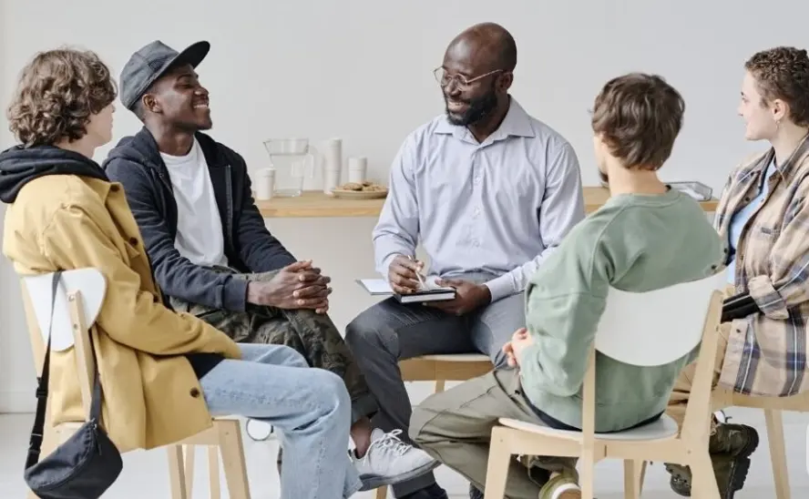 A facilitator taking notes while leading a small, smiling group seated in a circle of chairs